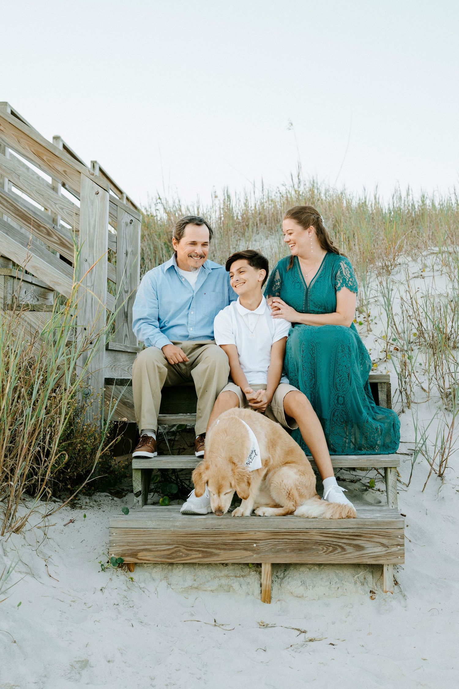A couple sits on wooden beach steps with their golden retriever, surrounded by sand dunes and sea grass.