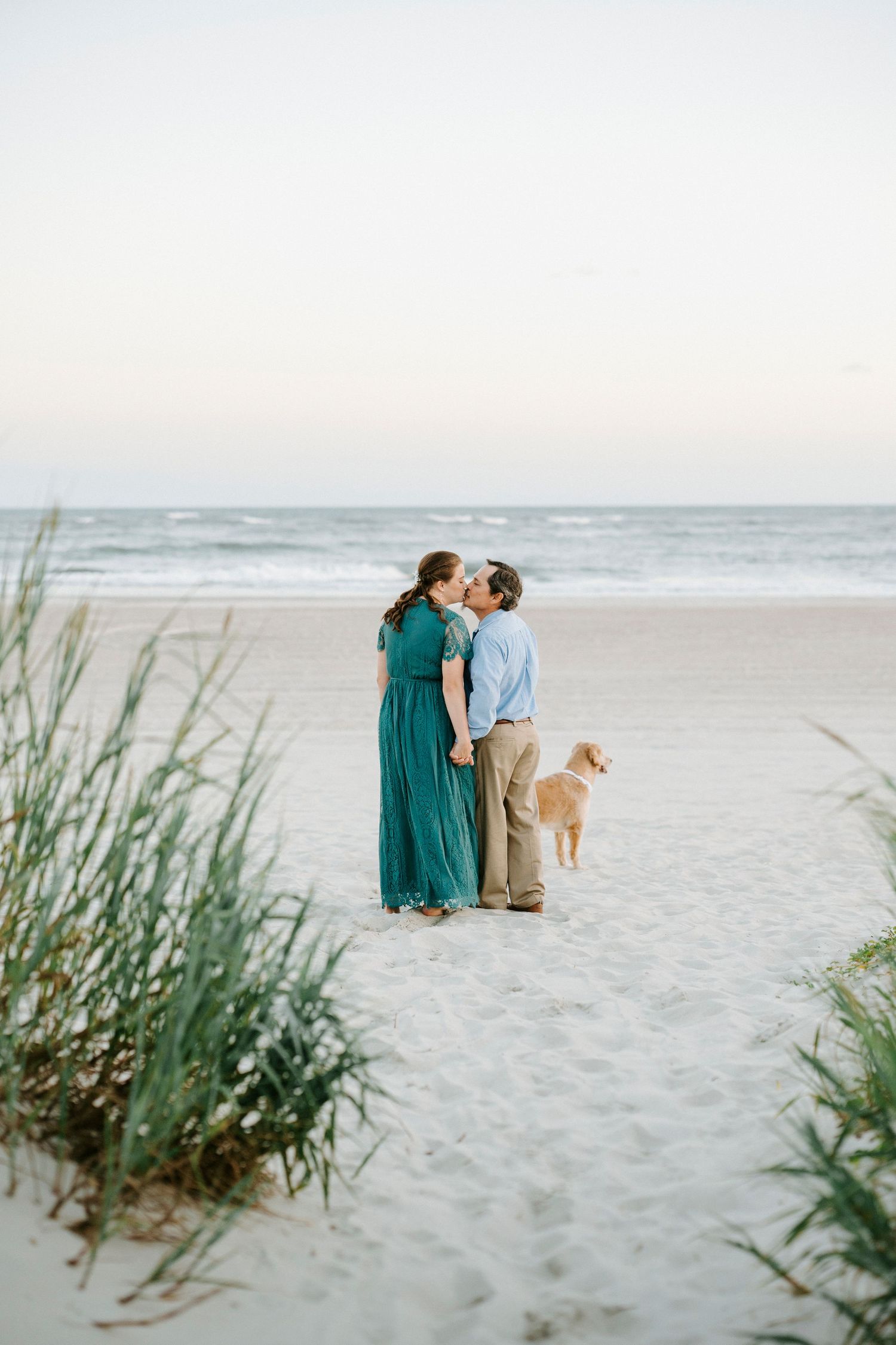 A couple walks along a serene beach with sea grass in the foreground during a peaceful sunset.