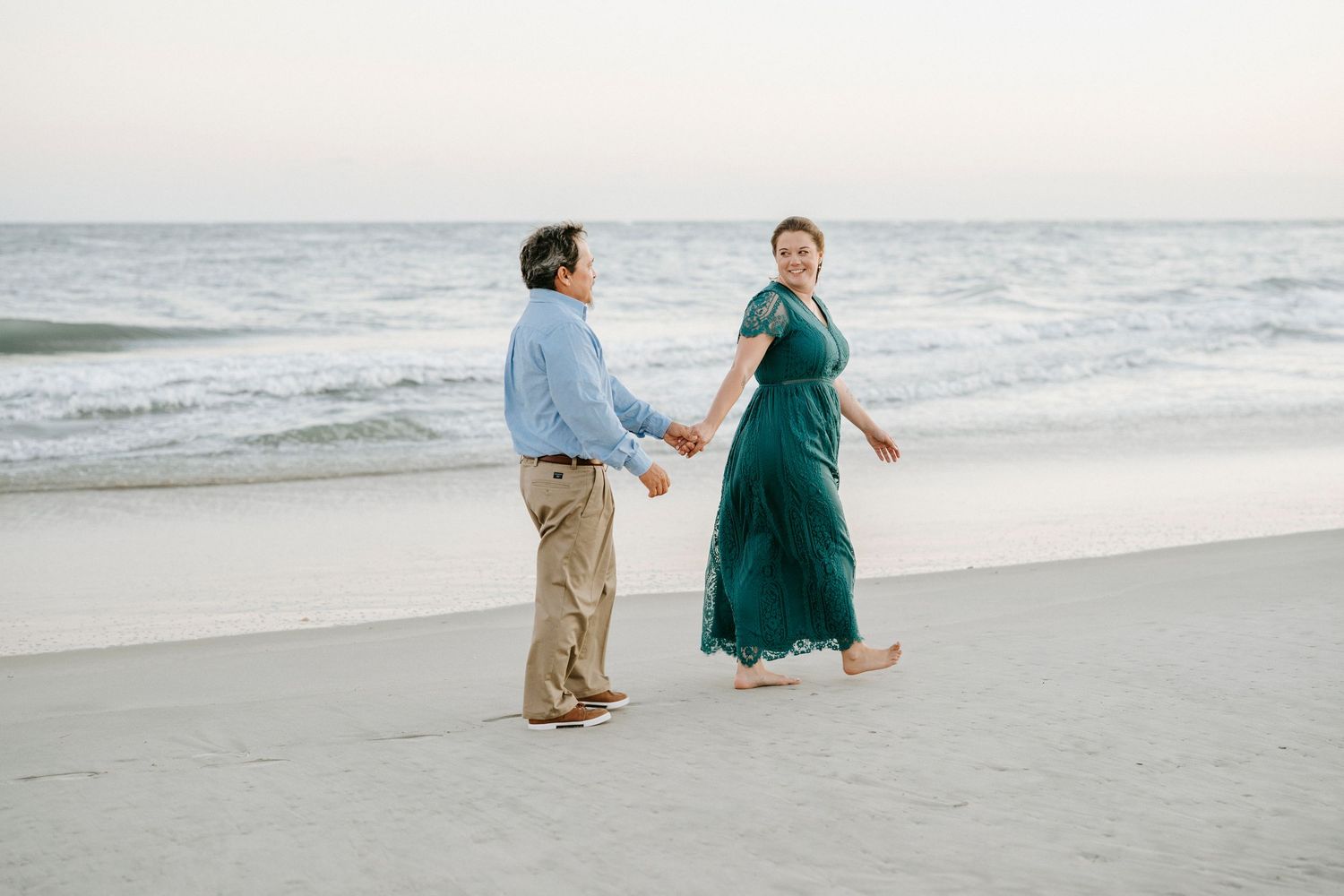 A couple walks hand in hand along a serene beach at dusk, with the woman wearing a flowing emerald green dress.