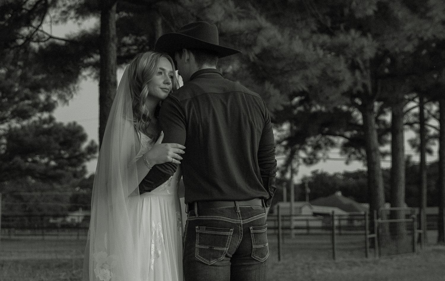 Black and white photo of a bride and groom sharing a romantic moment outdoors near trees at dusk.
