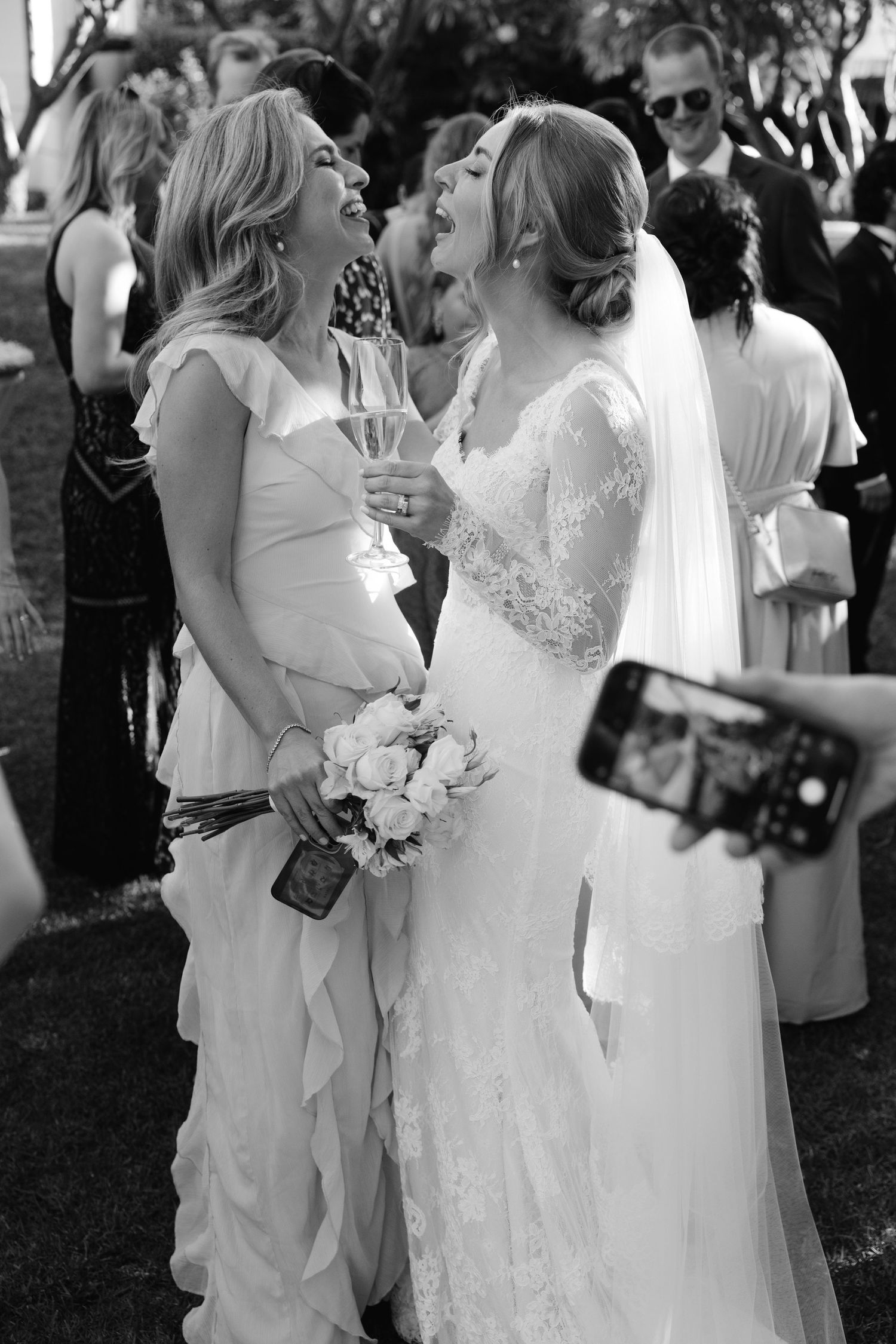 A joyful candid moment between a bride and bridesmaid sharing laughter at an outdoor wedding celebration.