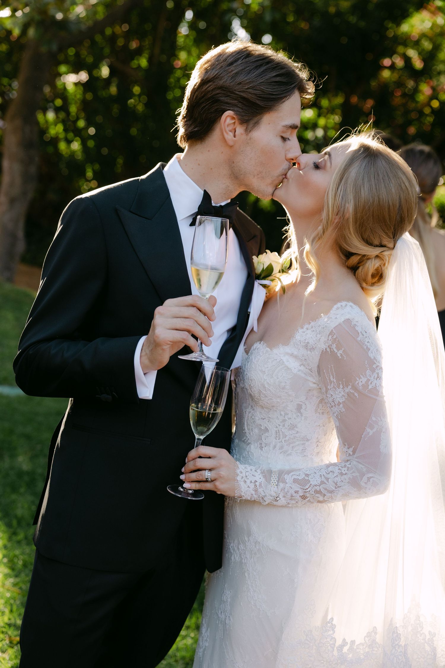Romantic newlyweds share champagne and kisses in an outdoor garden setting during golden hour.