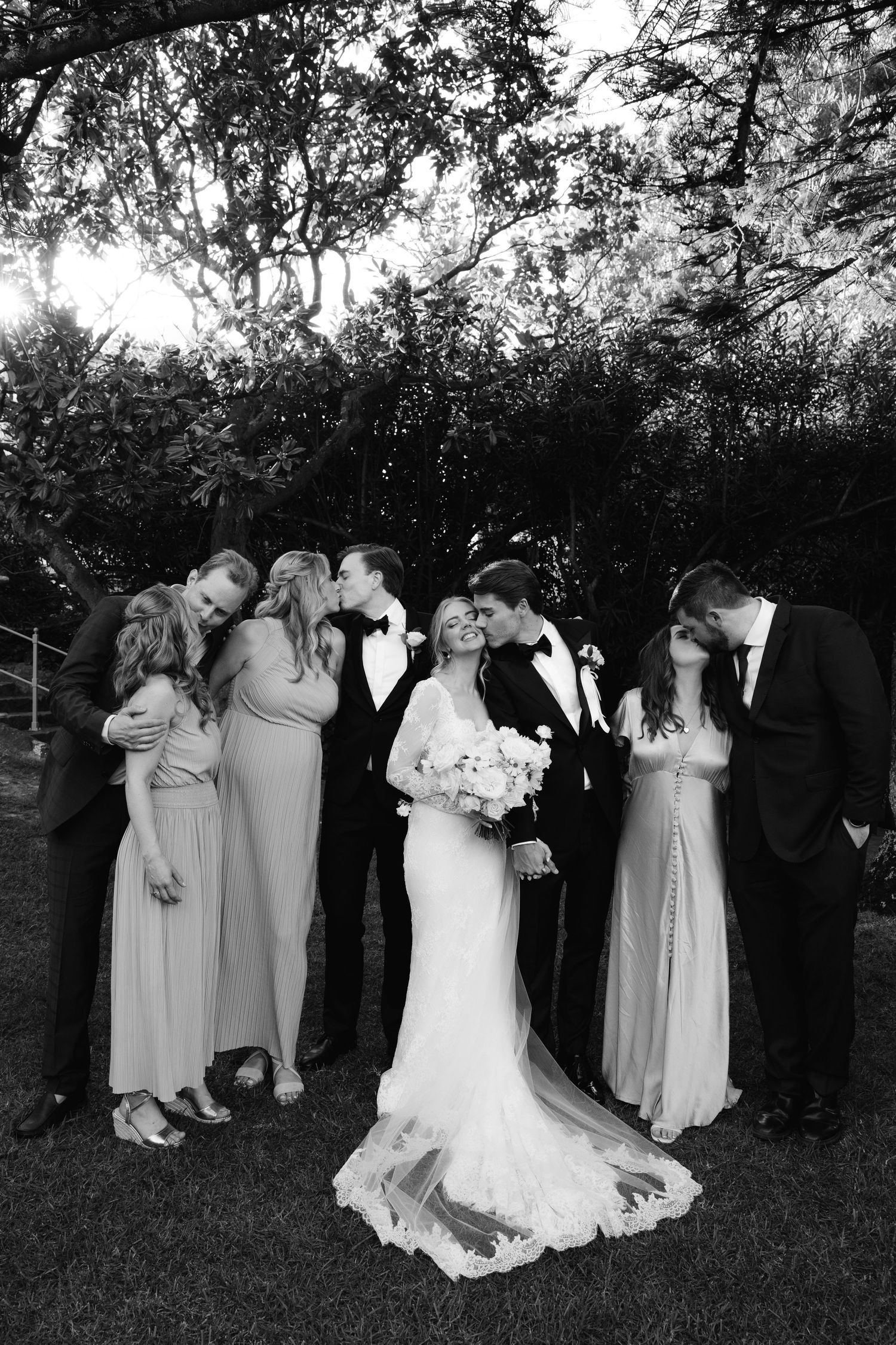 A wedding party in formal attire poses together outdoors at dusk in a garden setting.