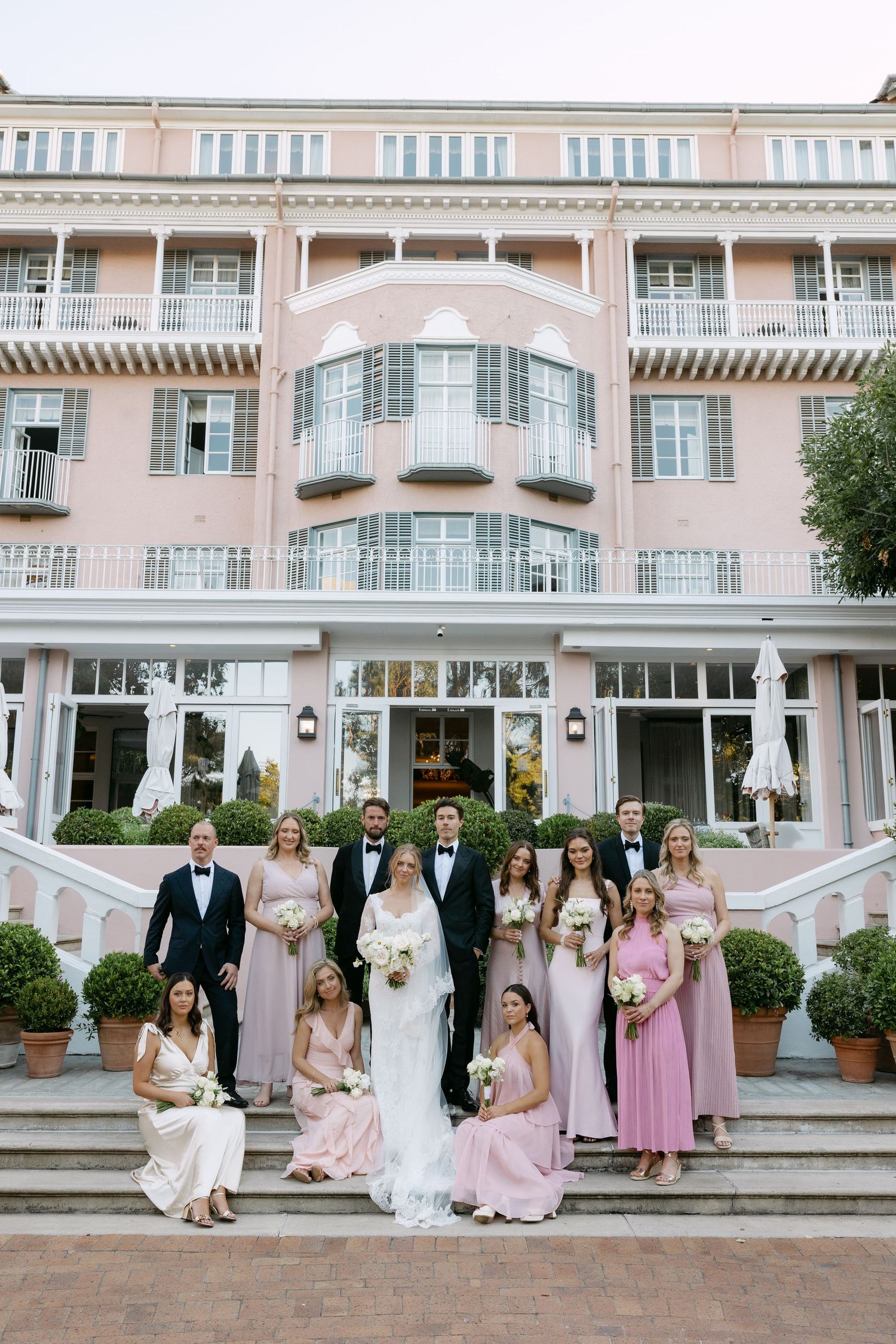 Wedding party poses on grand staircase of pink luxury hotel with classic architecture and elegant details.