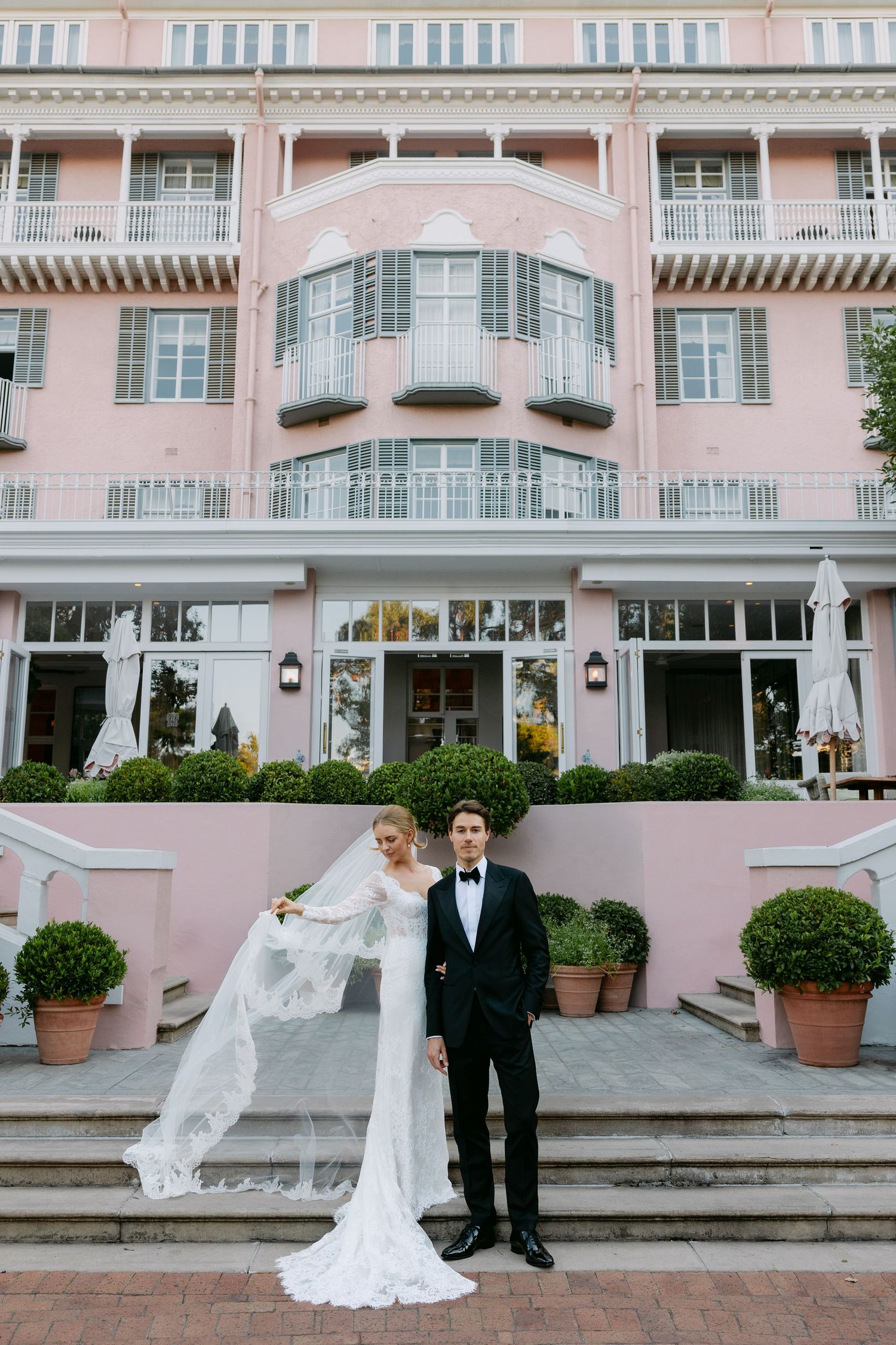 A wedding couple poses in front of a grand pink Mediterranean-style building with elegant archways and potted plants.