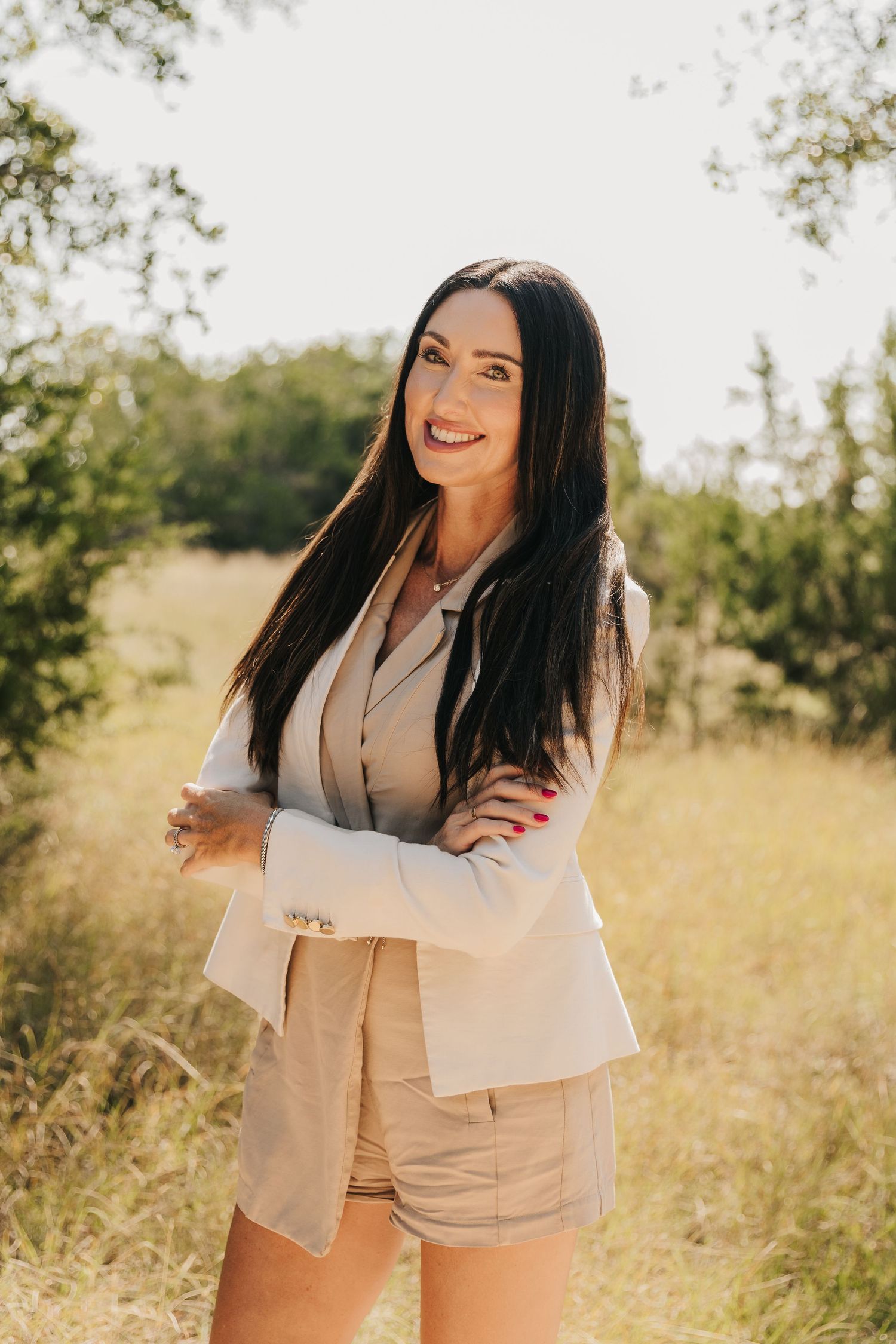 A person in a beige blazer and shorts stands smiling in a sunny field with trees in the background.