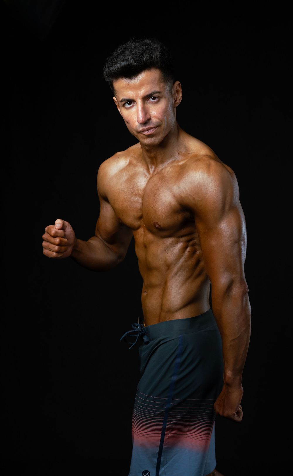 Athletic muscular physique of a fitness model posing shirtless against black background in gradient shorts.