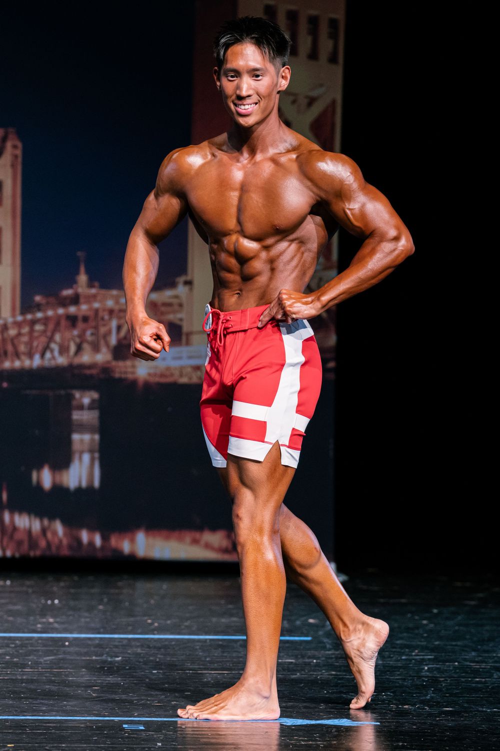 Male bodybuilder performs posing routine on stage wearing red shorts during competition.