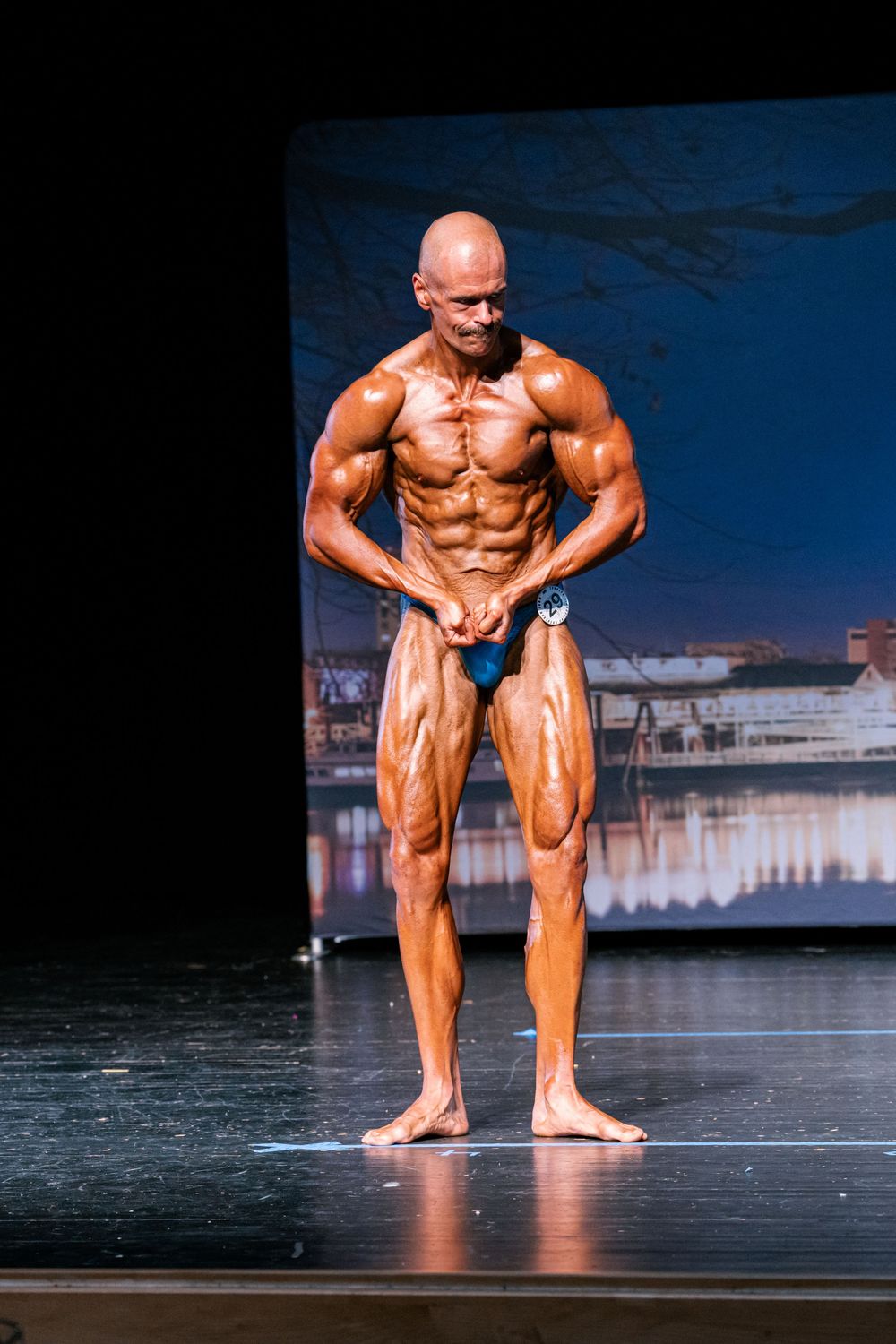 Muscular bodybuilder poses on stage during competition showing defined physique under bright lights.