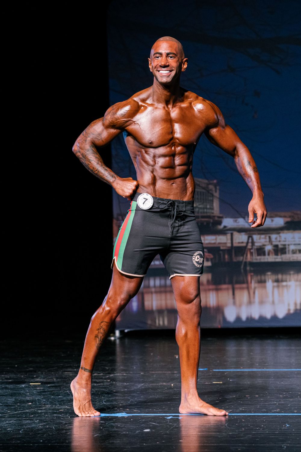 Muscular shirtless athlete in black shorts performs dynamic stretching poses on dark stage with dramatic lighting.