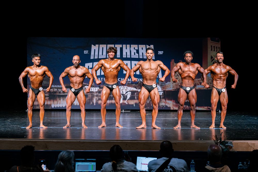 Bodybuilders in matching dark swimsuits pose on stage during a competitive fitness competition.