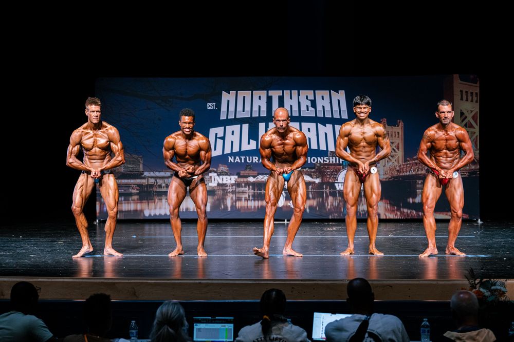Bodybuilding competitors pose on stage during a competition showing off their muscular physiques under bright lights.