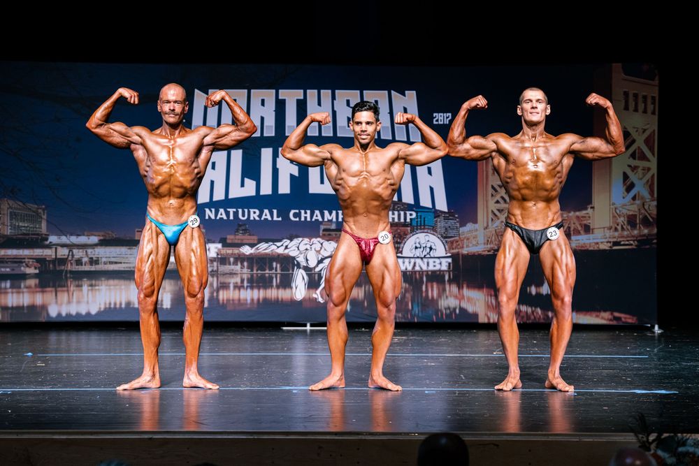 Bodybuilders strike poses on stage during a competitive bodybuilding show with blue lighting and backdrop.