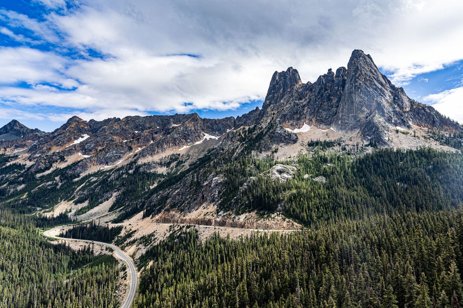 Highway winds through mountain pass with trees and rising rocky peaks under partly cloudy skies.