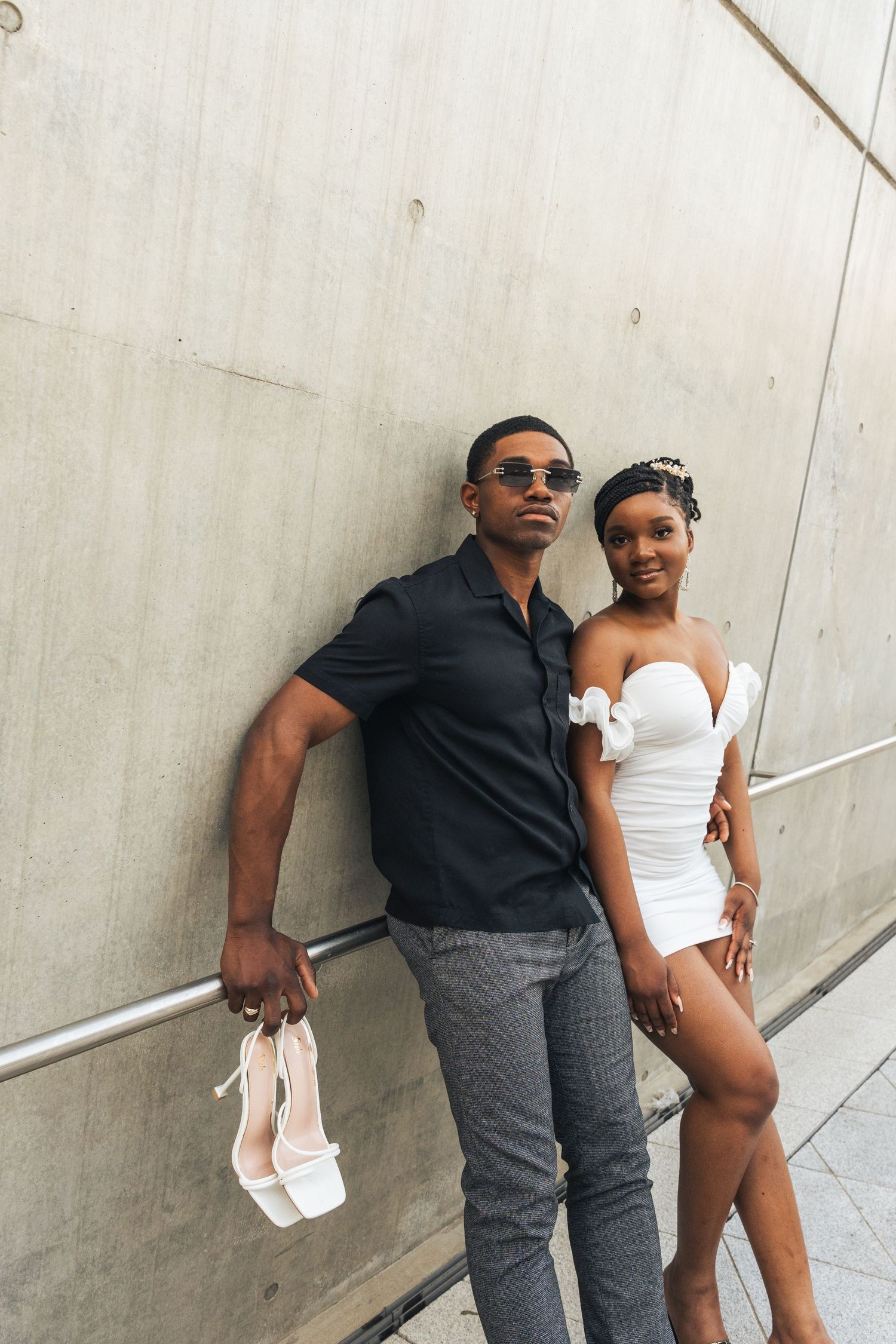 Two people in contrasting black and white outfits pose casually against a concrete wall in an indoor setting.