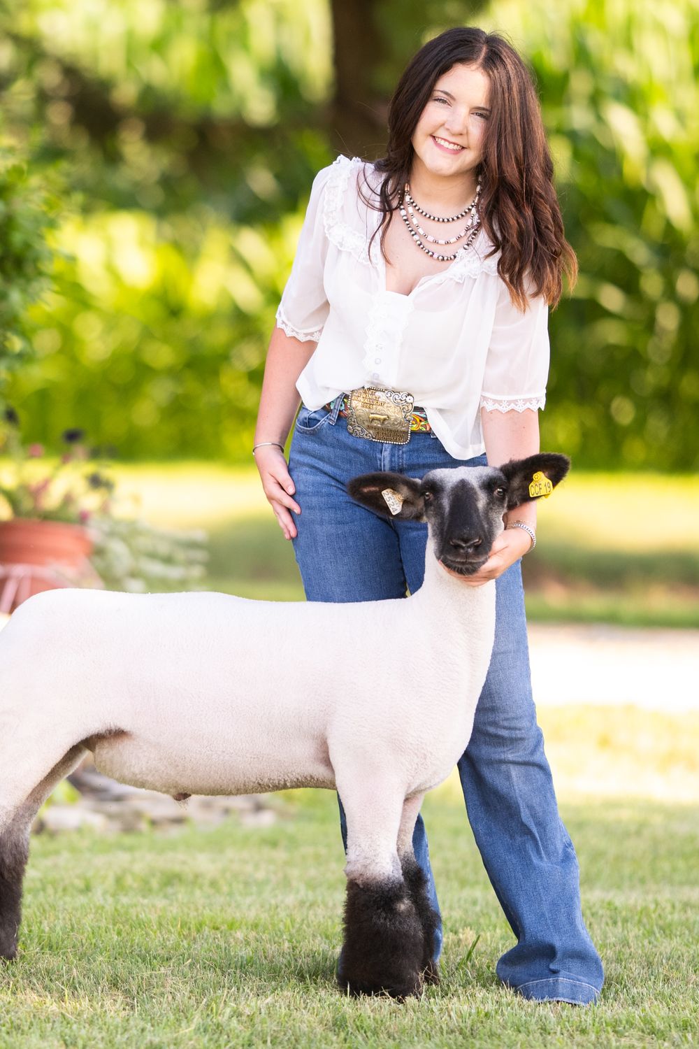 A show lamb poses with someone in a white top and jeans in an outdoor setting with green foliage.