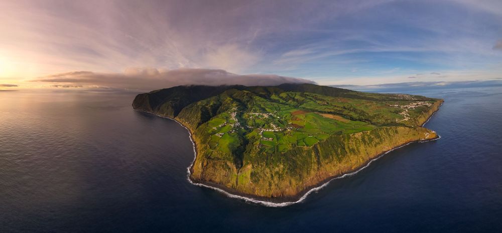 Aerial panoramic view of a rugged coastline with green cliffs and ocean waves during sunset.