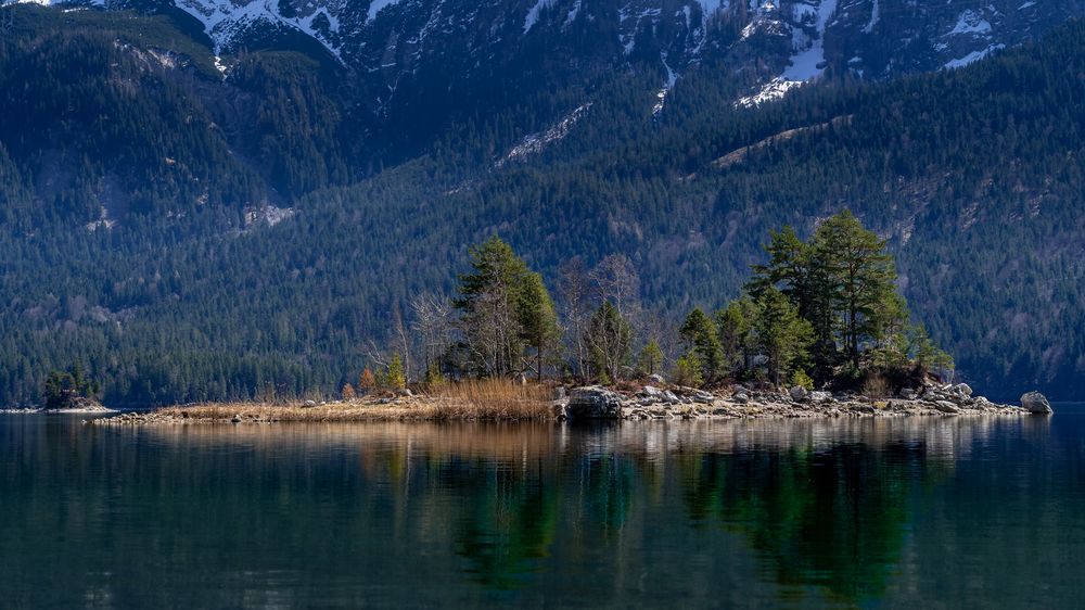 Snow-capped mountain range reflecting in calm lake waters with evergreen forest in foreground.
