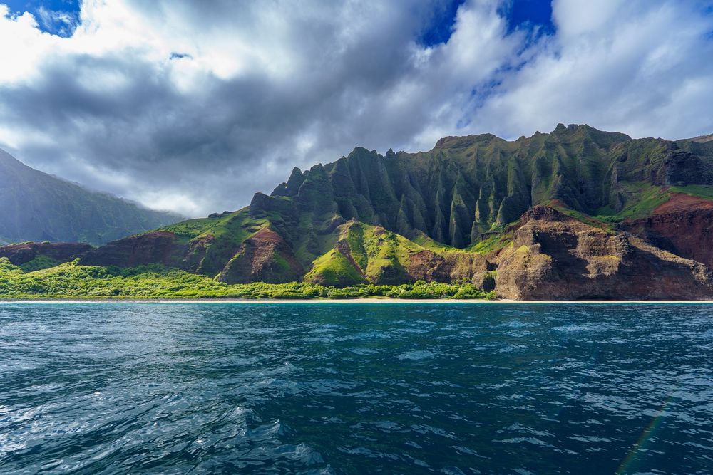 Panoramic view of the rugged Na Pali coast of Kauai with dramatic cliffs rising from turquoise waters under cloudy skies.