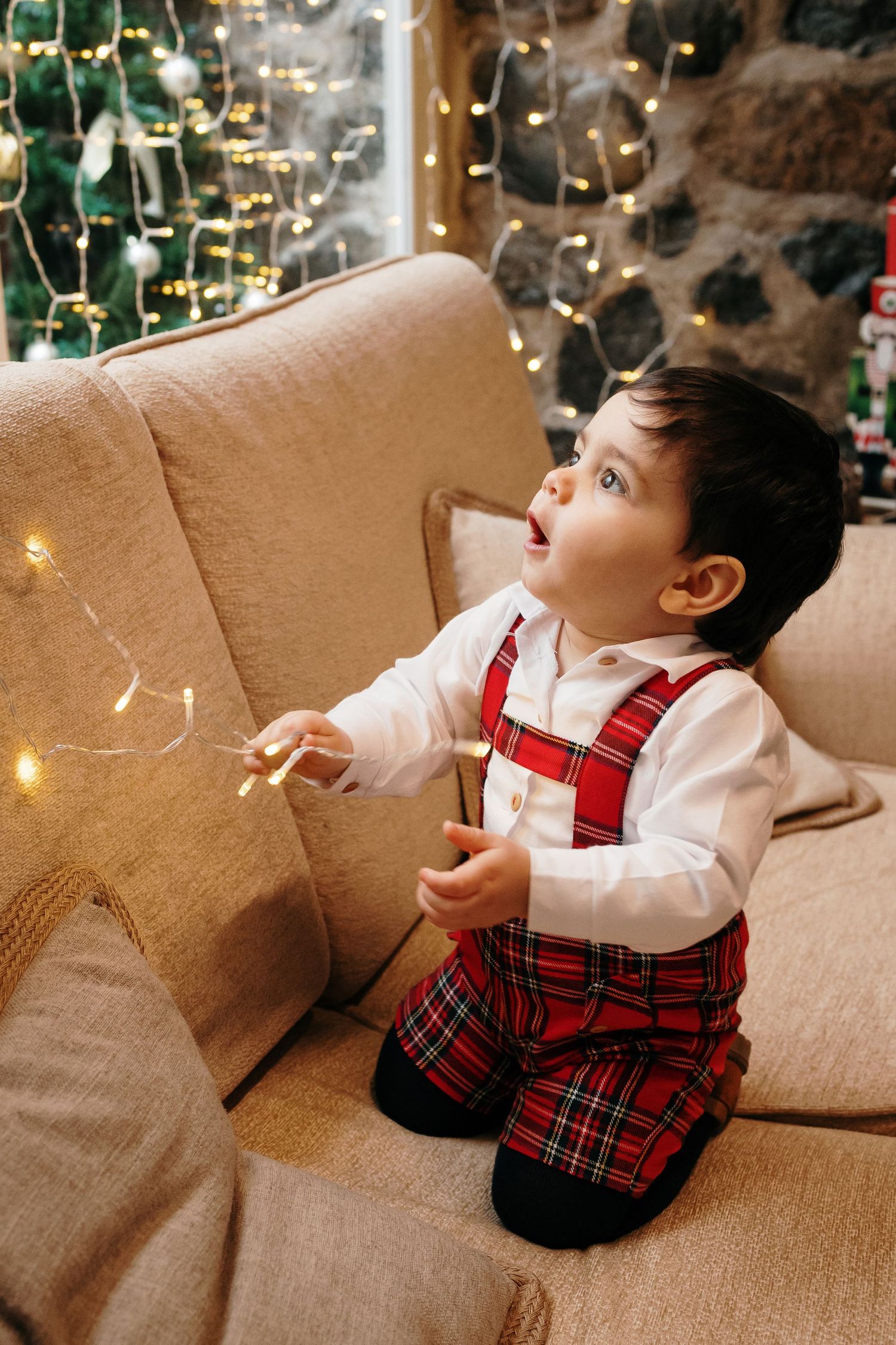Child in red plaid outfit sits on beige couch near Christmas decorations.