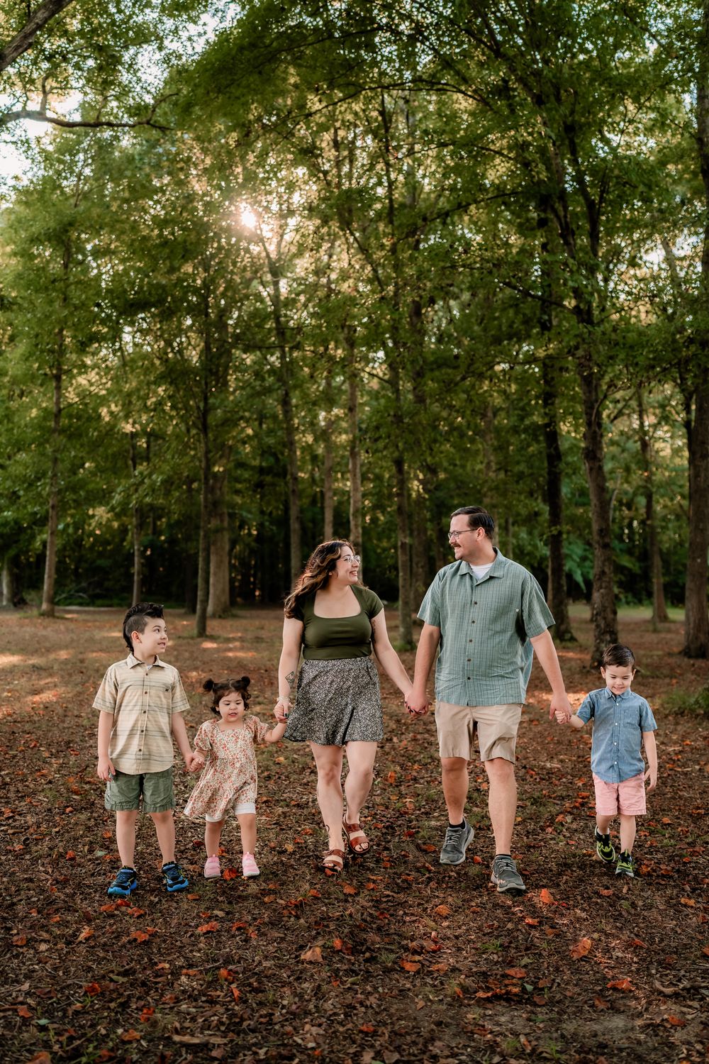 A family walking together through a wooded park with fallen autumn leaves covering the ground.
