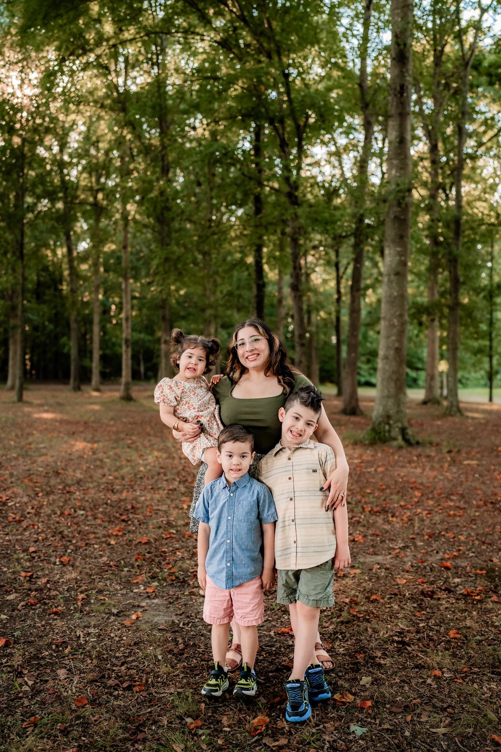 Three children playing and laughing together in a wooded park area covered with fallen autumn leaves.