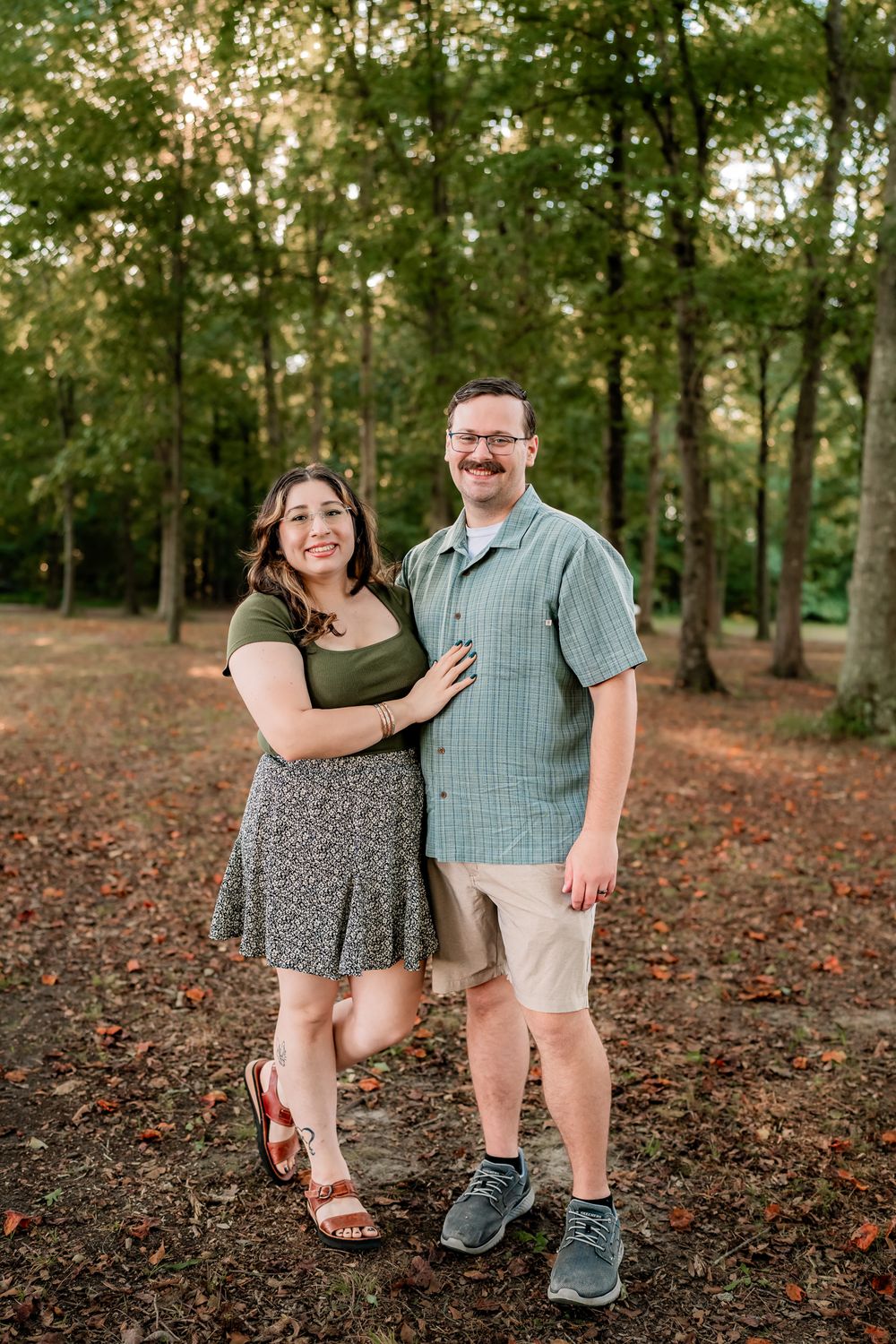 A couple poses playfully in a wooded area with fallen autumn leaves on the ground during golden hour.