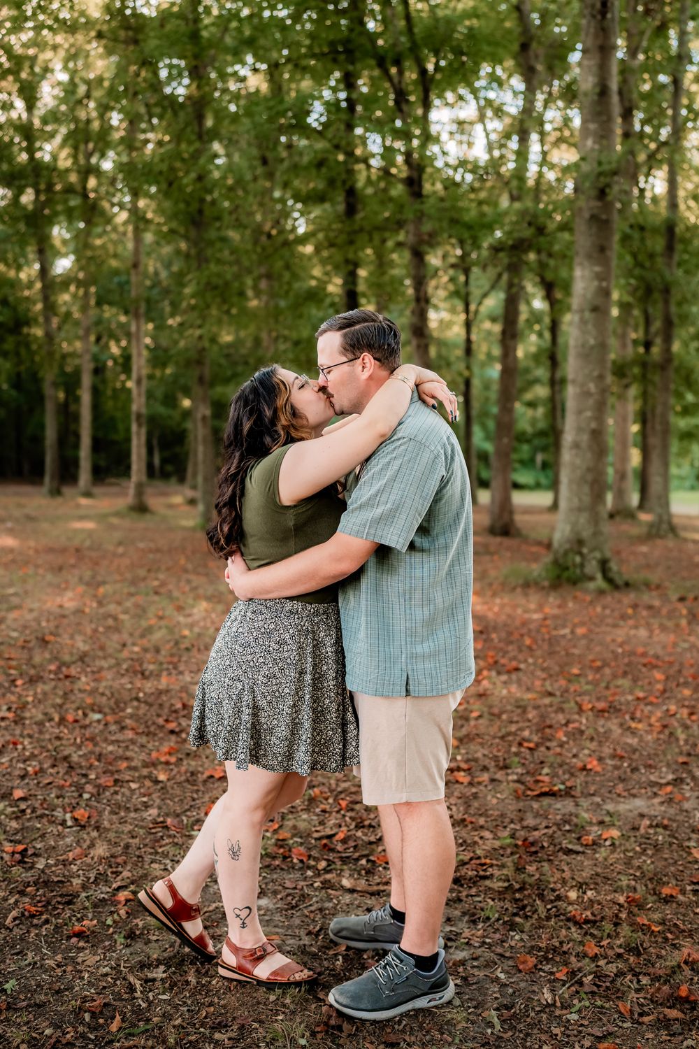 A couple embraces in a wooded area with fallen autumn leaves covering the ground.
