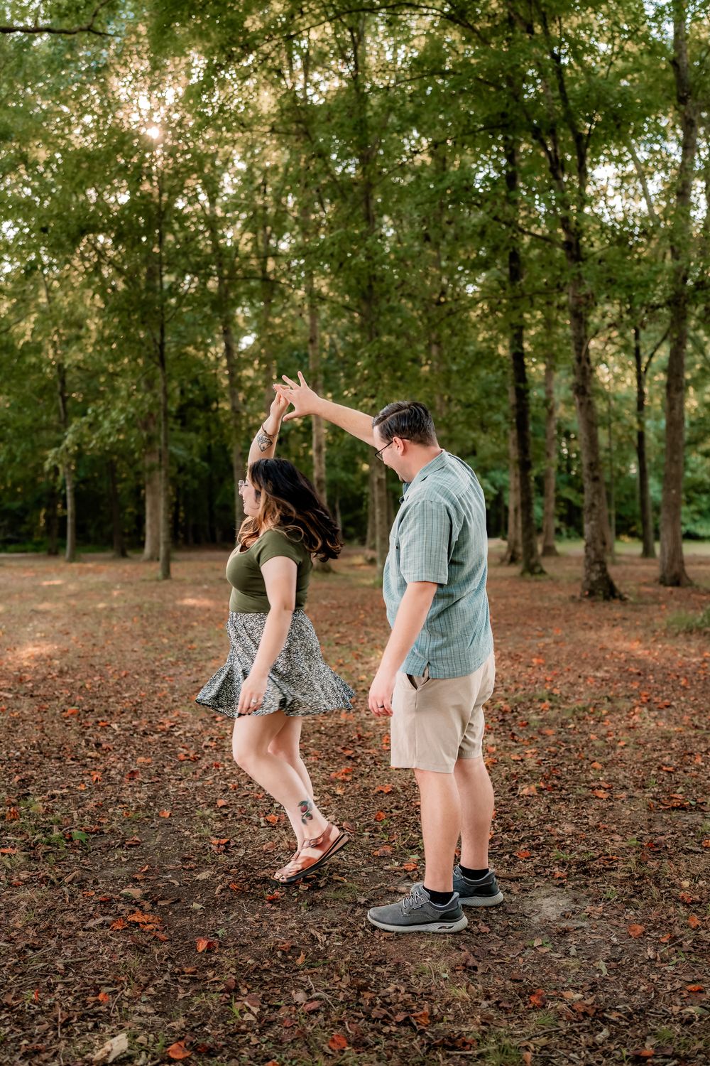 A couple playfully dances together on fallen autumn leaves in a wooded park setting during golden hour.