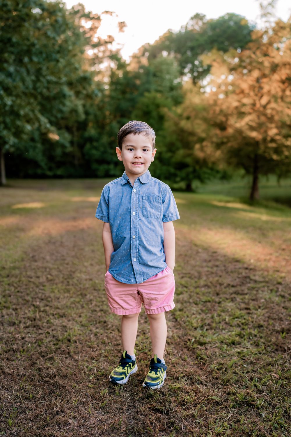 A young child stands in a park wearing a blue button-down shirt and pink shorts on a warm autumn afternoon.