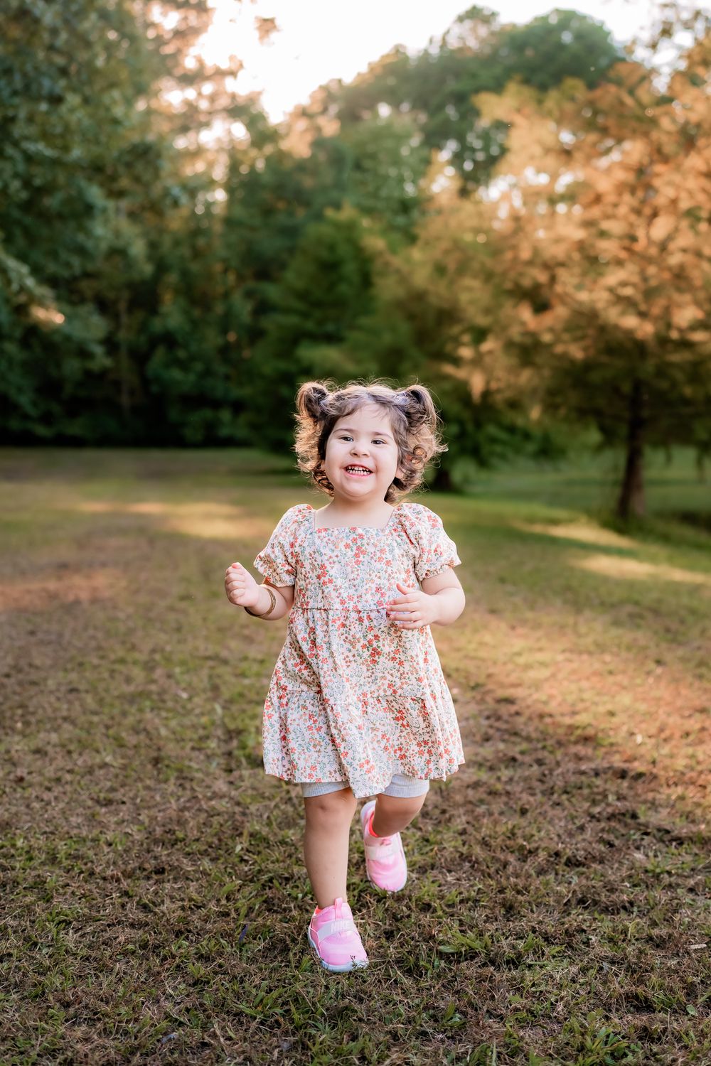 A toddler in a floral dress and pink shoes runs joyfully through a park at golden hour.