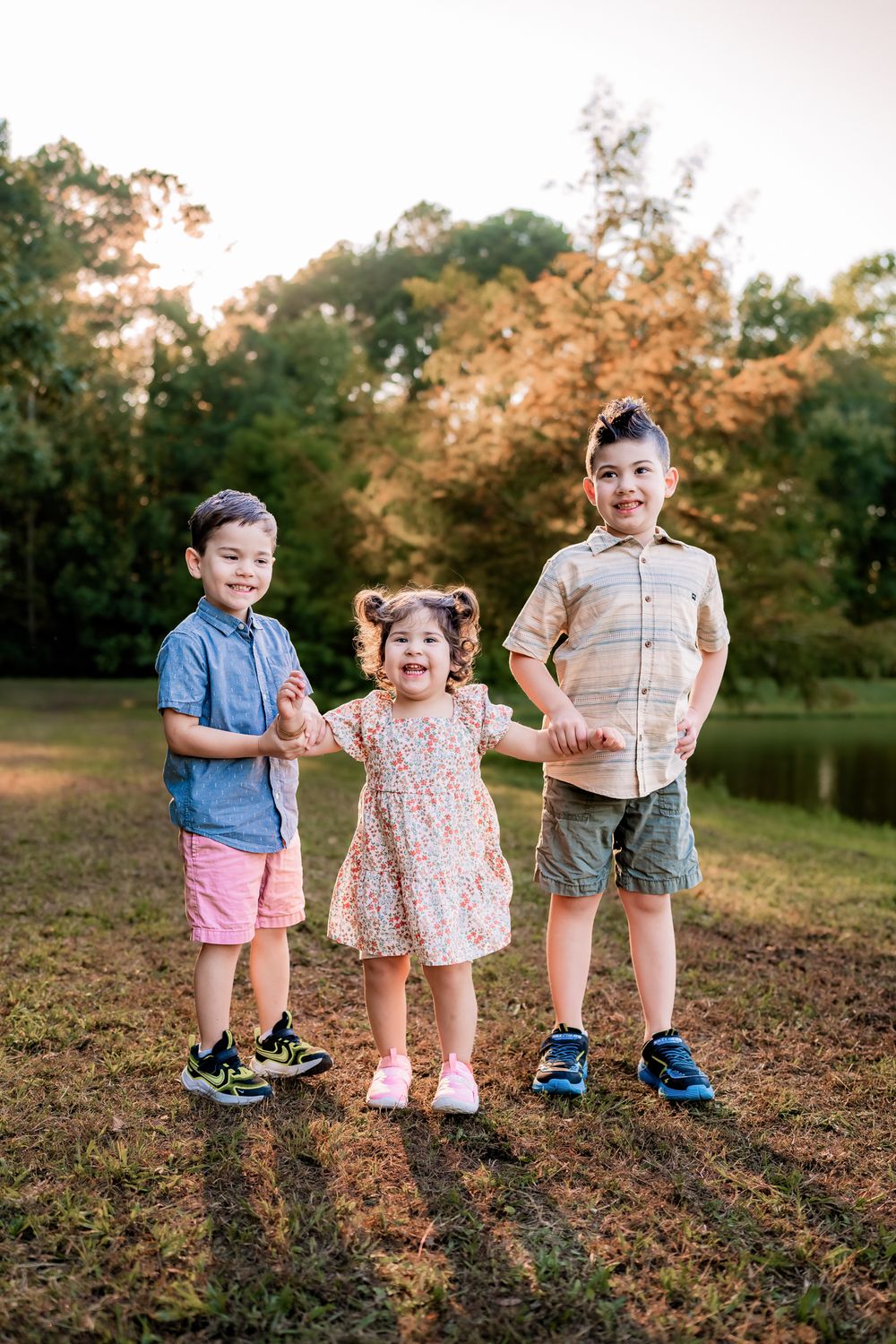 Three young children stand together smiling by a pond on an autumn evening.