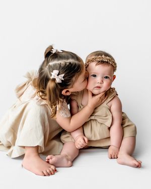 Two young children in cream-colored outfits share sweet moments together against a white background.