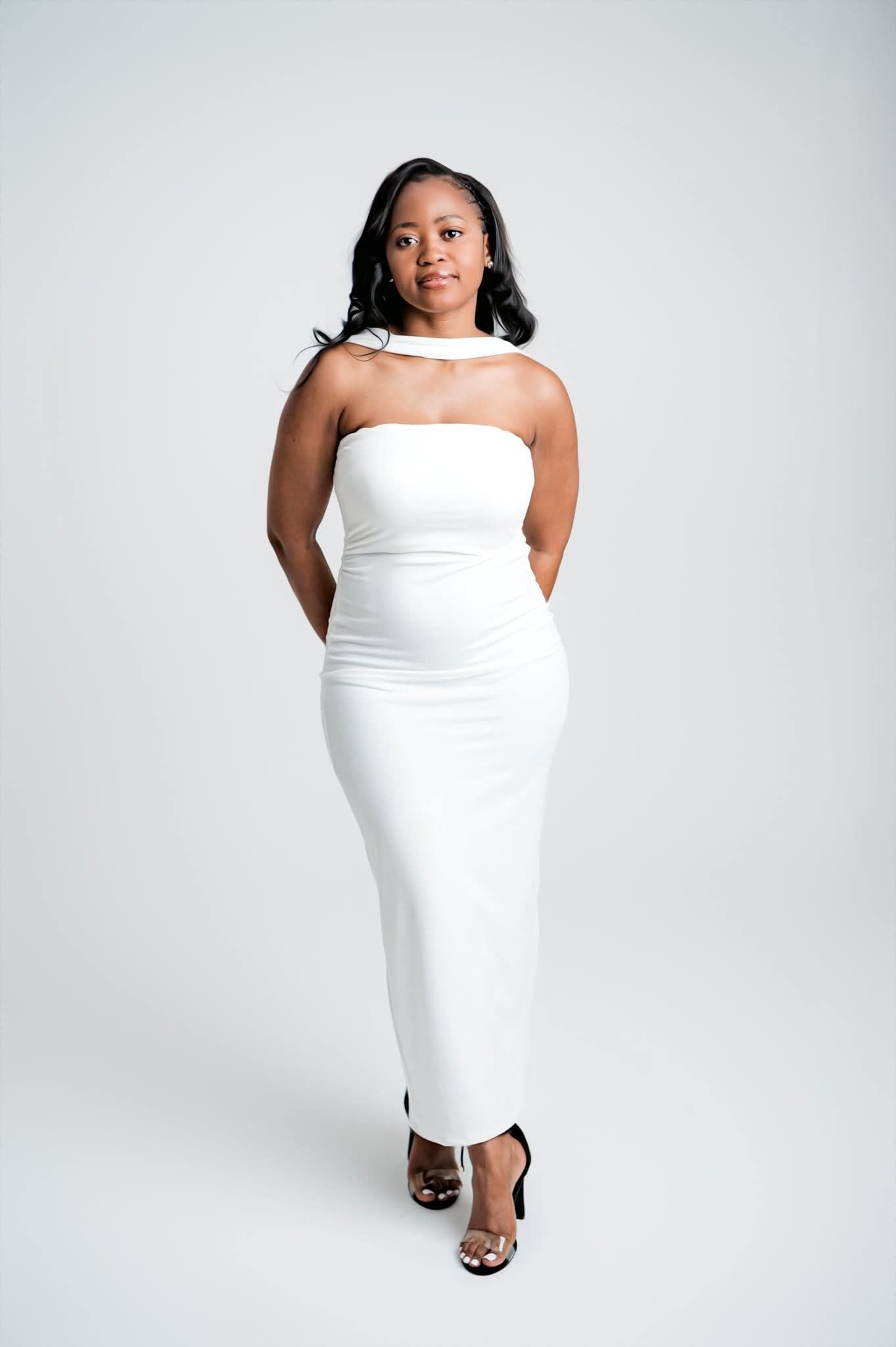 A person poses in a form-fitting white strapless dress and clear heels against a light gray studio background.