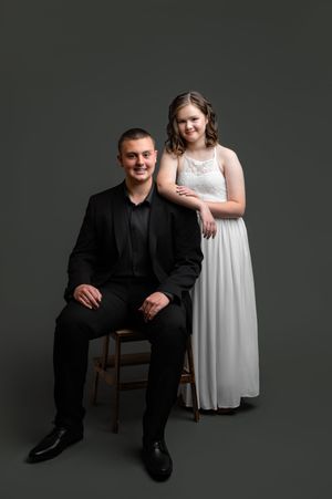Studio portrait with dark background showing two people in black and white formal attire posing together.