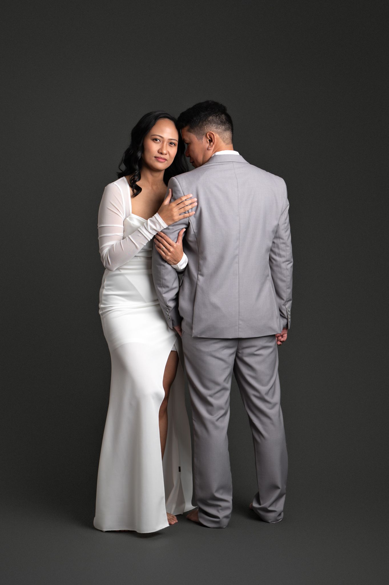 Couple in elegant wedding attire, bride in white gown and groom in gray suit, posing romantically in studio.