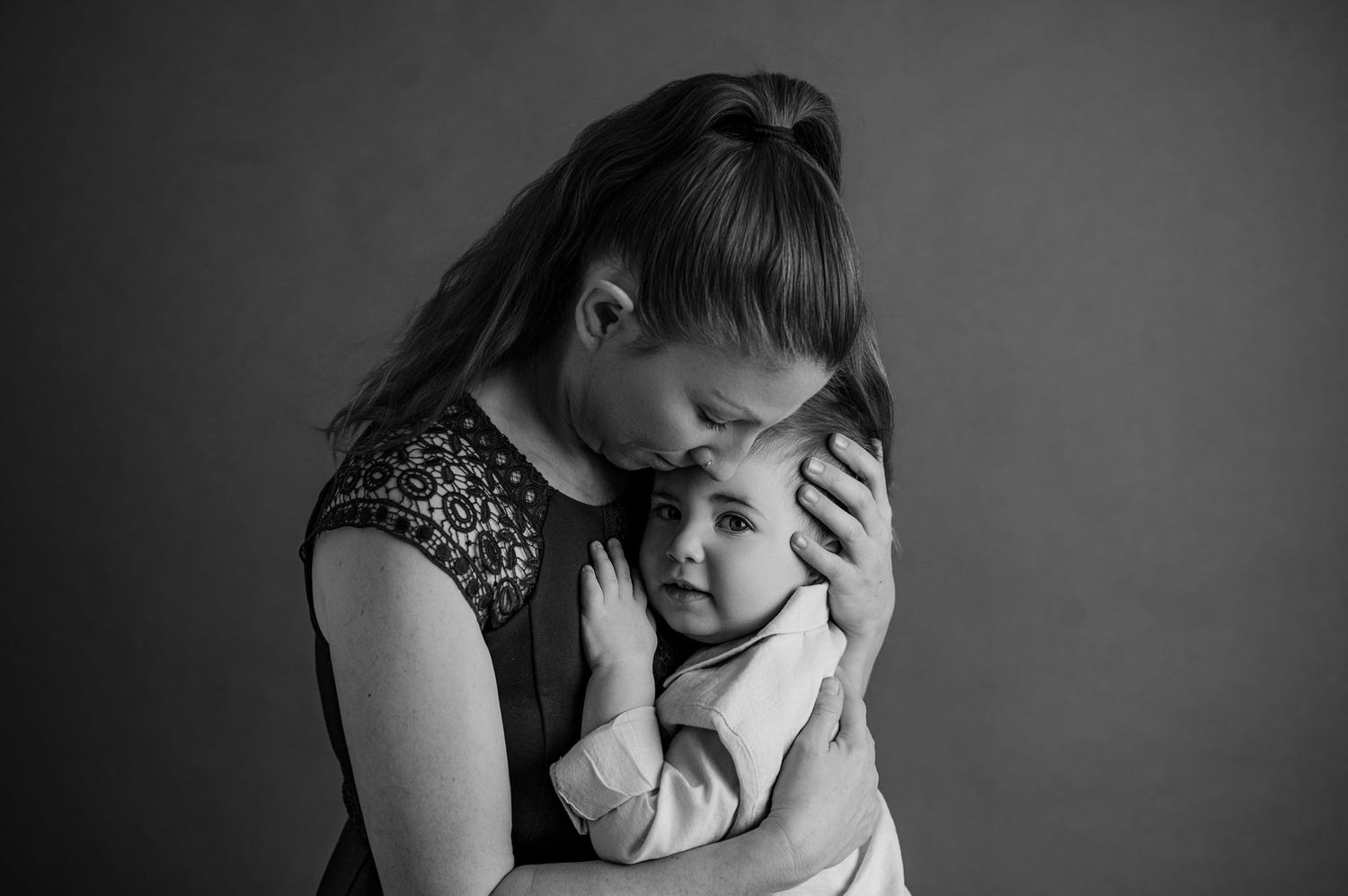 Black and white portrait showing tender embrace between parent and baby in intimate studio setting.