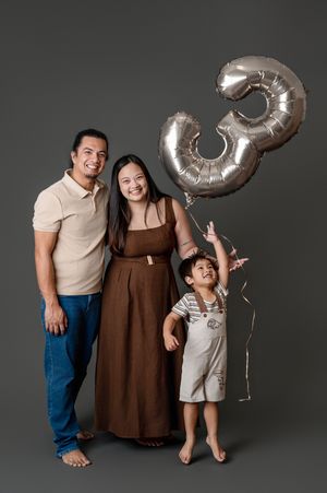 Family of three posing with a silver number 3 balloon, celebrating a third birthday in a studio.