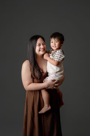 A smiling woman in a brown dress holds a happy toddler boy in overalls against a gray studio background.