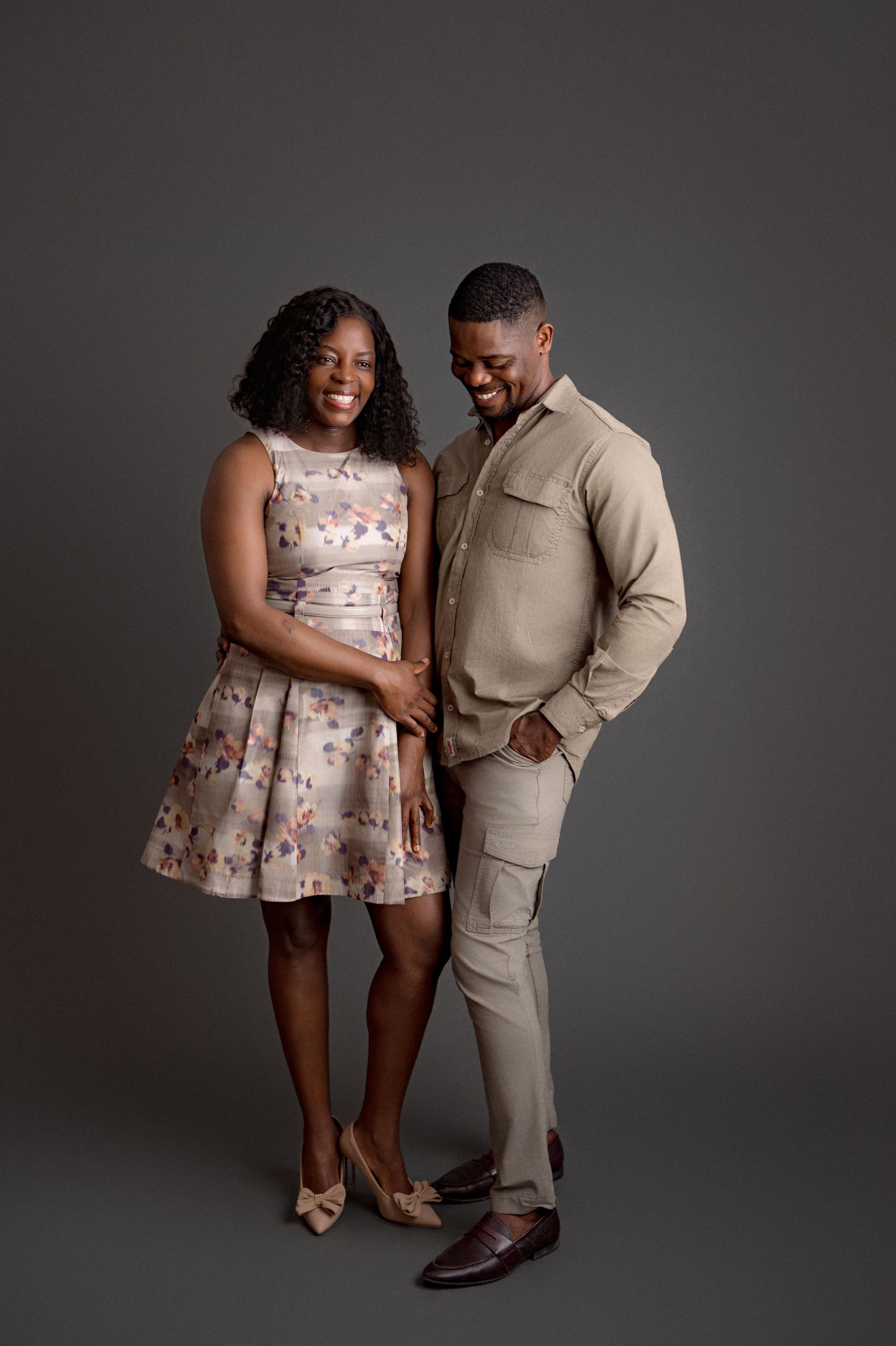 A couple poses together against a gray backdrop wearing coordinated beige and floral formal attire.