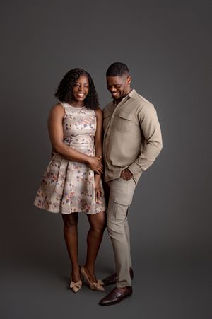 A couple poses together against a gray backdrop wearing coordinated beige and floral formal attire.