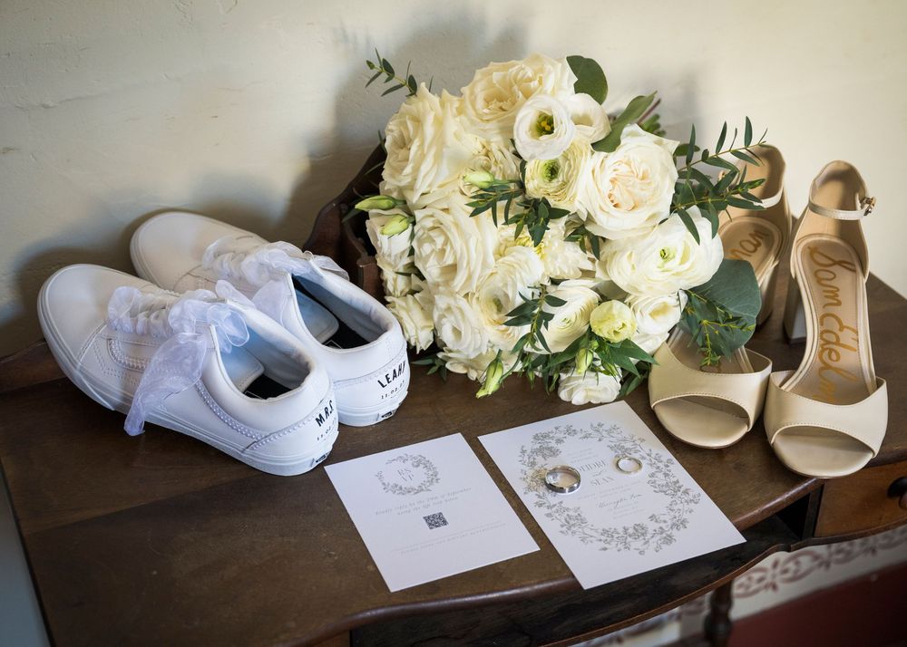Wedding accessories including white shoes, floral bouquets, and invitation cards arranged on a dark surface.
