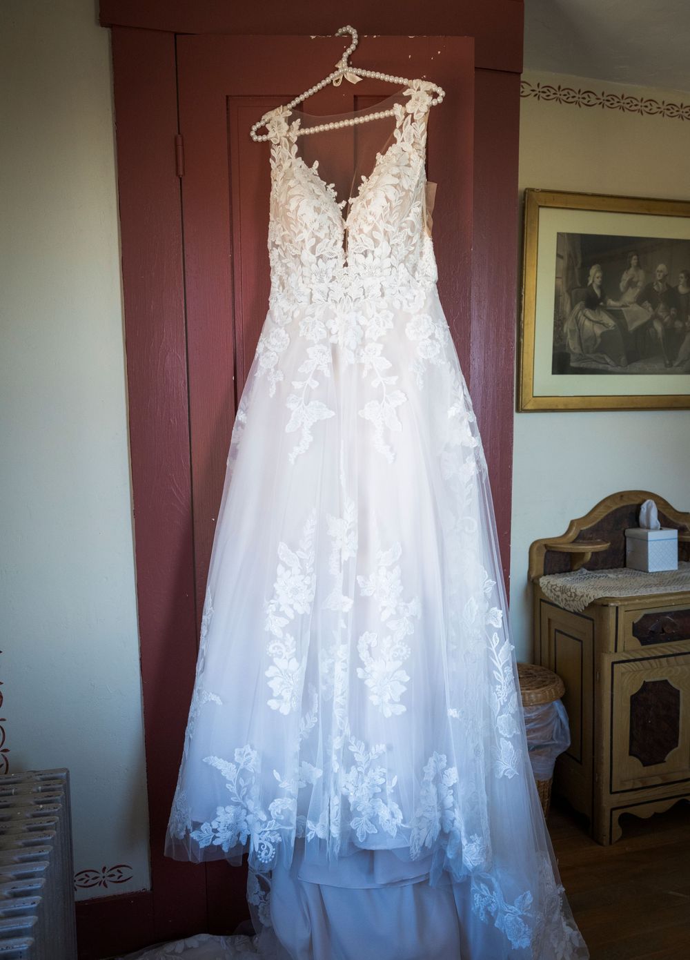 White lace wedding dress with floral details hanging on display against burgundy wall.