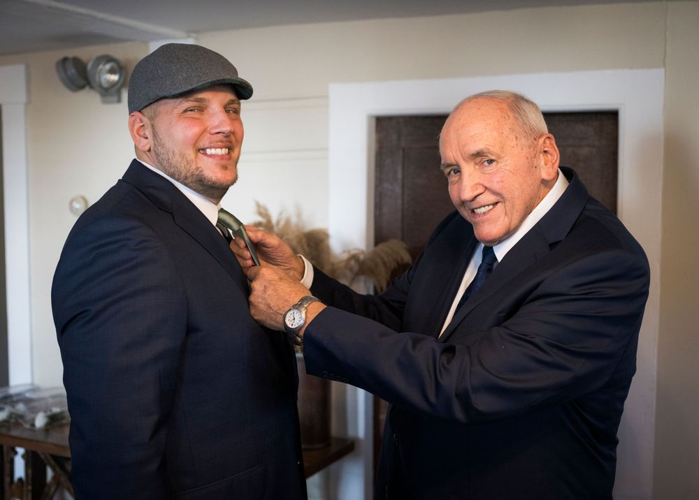 Groomsmen in black suits and baseball caps share laughs while getting ready for a wedding celebration.