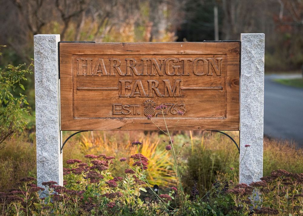 Wooden Harrington Farm entrance sign mounted between white posts with fall foliage in background.