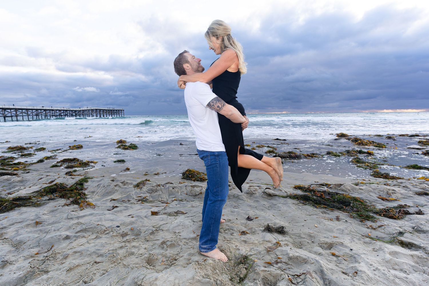 San Diego couple portrait photography of a man lifting a woman on a sandy beach with a wooden pier in the background.