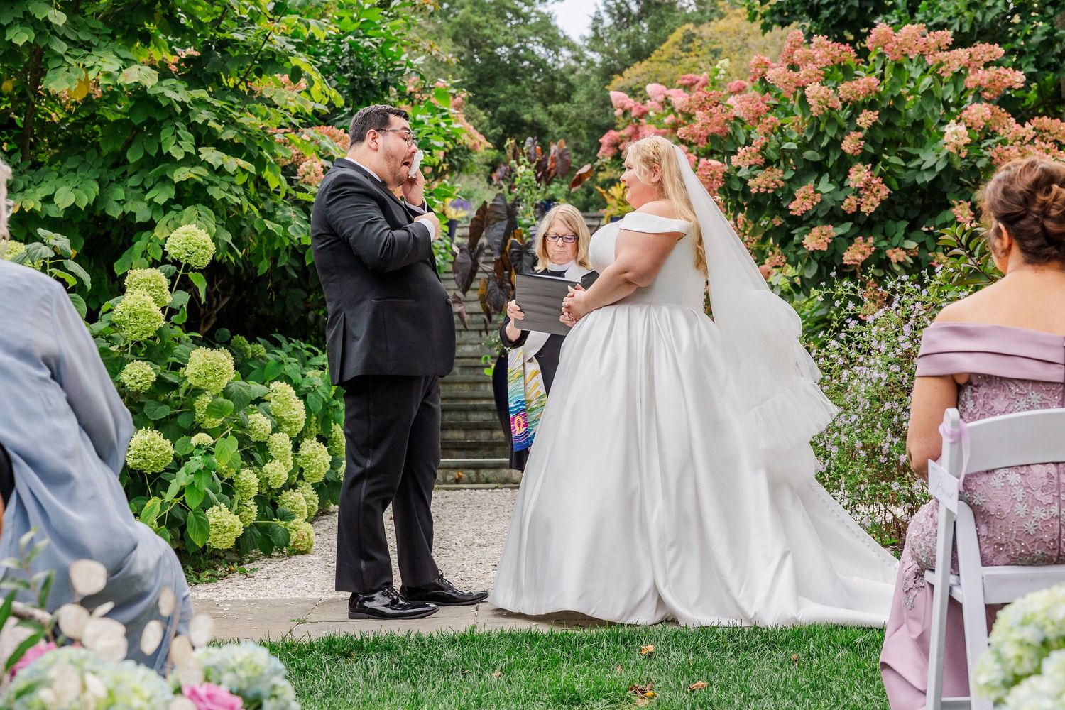 Wedding ceremony taking place in a lush garden setting with blooming hydrangeas and coral flowers in the background.