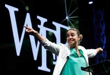 A keynote speaker in a bright green blazer gestures expressively on stage during a dynamic performance.