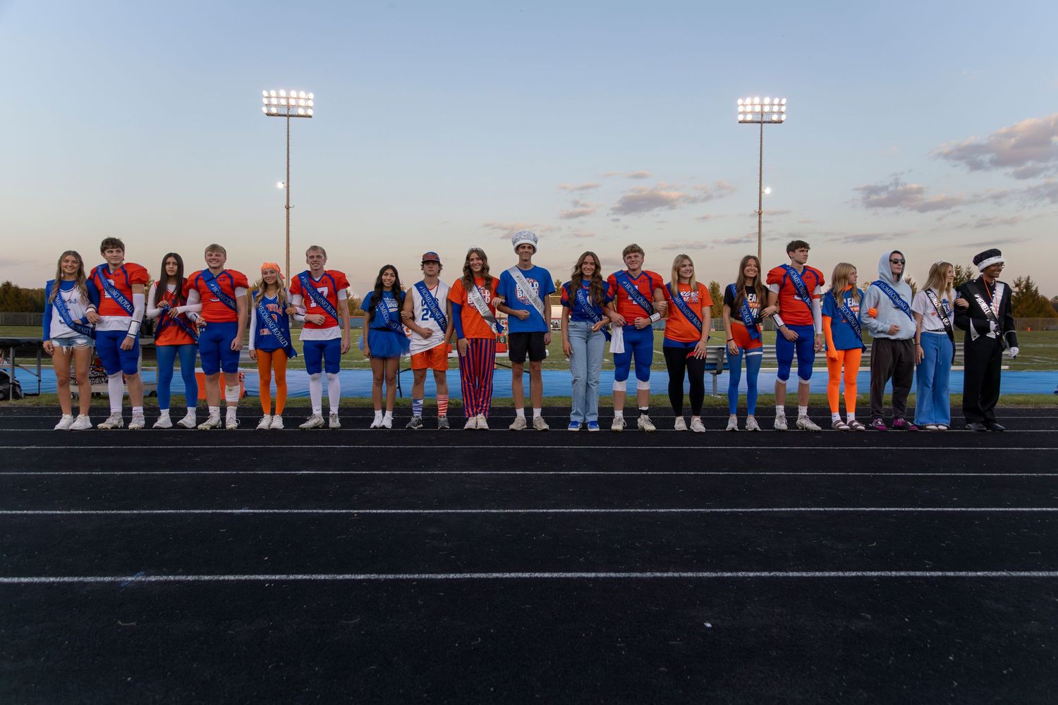 Sports team members line up on track field at dusk under stadium lights.