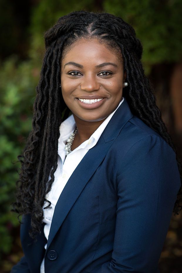 Professional woman in navy blazer with locs hairstyle smiling outdoors in a corporate headshot photo.